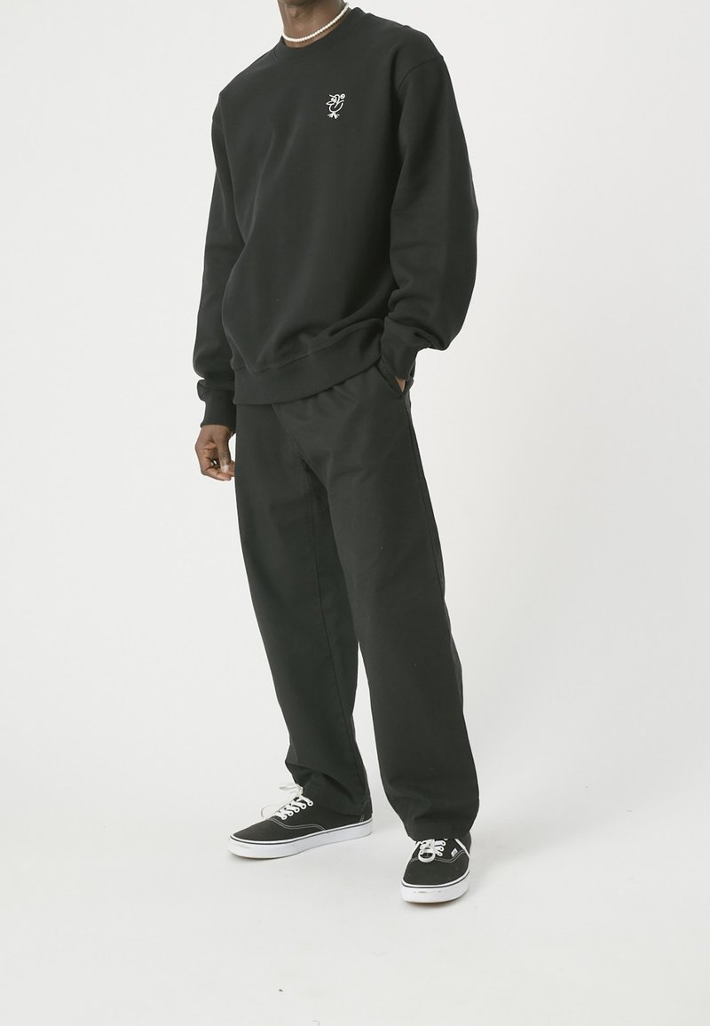 Sudadera negra con un pequeño logo blanco en el pecho, combinada con pantalones negros de corte holgado y zapatillas negras con suelas blancas.