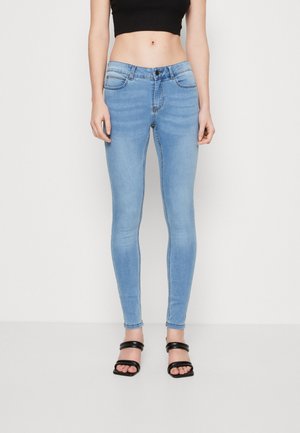 Femme portant un jean skinny bleu clair, un top court noir et des sandales à talons noires, debout devant un fond blanc uni.