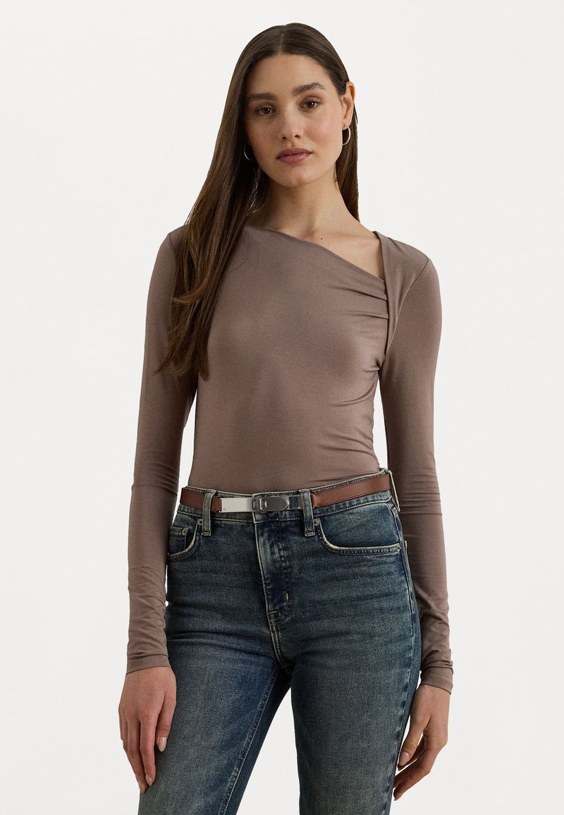 Brauner Langarm-Top mit asymmetrischem Ausschnitt, kombiniert mit blauen Denim-Jeans und einem braunen Gürtel. Weicher, dehnbarer Stoff mit einer figurbetonten Silhouette.