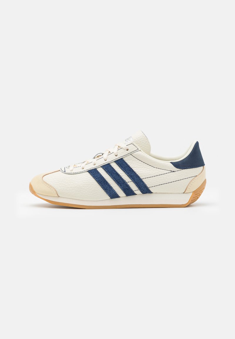 Zapatilla blanca de cuero con acentos en azul marino, que presenta tres rayas, una superficie texturizada y una suela de goma.