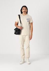 Beige t-shirt med blå logo, cremefarvede bukser, sort crossbody taske og hvide sneakers. Simpelt design, almindelig pasform med korte ærmer.