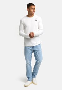 Camiseta blanca de manga larga con cuello redondo, que presenta un pequeño logo negro en el pecho, combinada con jeans de color azul claro y zapatillas blancas.