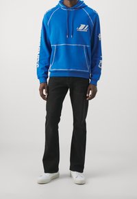 Homme portant un sweat à capuche bleu avec des motifs blancs, un pantalon noir et des baskets blanches, debout devant un fond clair uni.