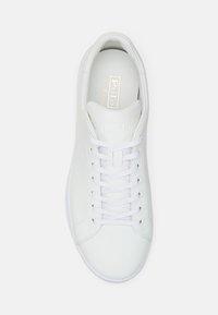 Bílé kožené tenisky s hladkou texturou, plochými tkaničkami, zaoblenou špičkou a zlatým logem na vnitřní stélce. Minimalistický design.