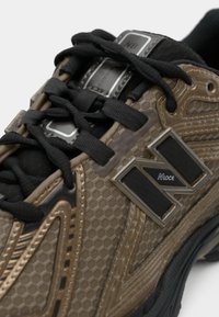 Brown sneaker with a textured fabric upper, black laces, metal accents, and a prominent raised logo on the side.