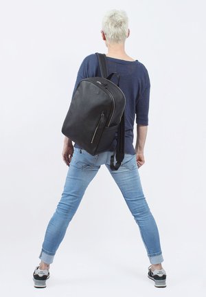 Zaino in pelle nera con texture liscia, dotato di una tasca frontale con zip e spallacci regolabili, indossato sulla schiena di una persona in abbigliamento casual.