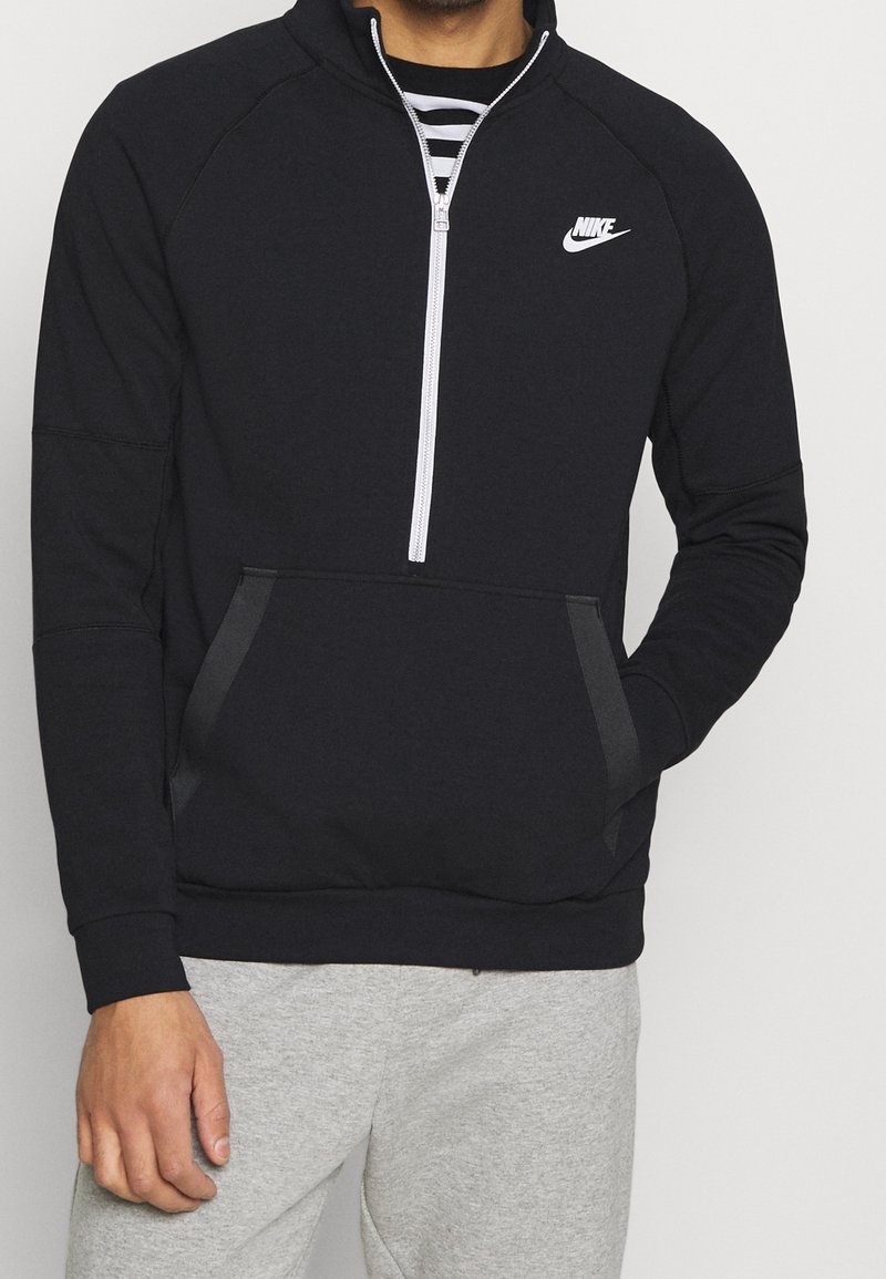 Muž v černé mikině s krátkým zipem značky Nike přes pruhované tričko, v šedých teplácích, s rukou v kapse.