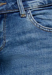Gros plan d'un jean en denim bleu montrant la poche avant, la boucle de ceinture et les coutures détaillées sur la texture du tissu.