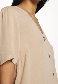 Kurzärmelige, blassbeige Bluse mit V-Ausschnitt, versehen mit einer texturierten Oberfläche und drei goldenen Knöpfen vorne.