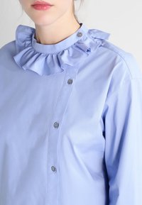 Camicia azzurra chiara con colletto arricciato, chiusura con bottone a pressione e tessuto liscio. Il design è strutturato con accenti di cucitura visibili.