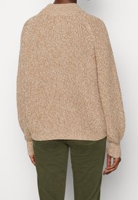 Beige pletený svetr s texturovaným povrchem a vysokým límcem. Má volné rukávy a uvolněný střih, kombinovaný se zelenými olivovými kalhotami.