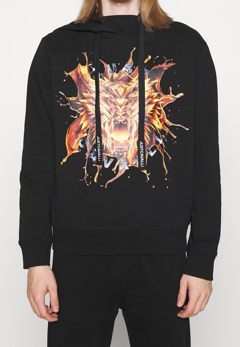 Sweat à capuche noir avec un graphisme de tête de lion géométrique enflammée et des lettres métalliques, porté par une personne visible des épaules aux hanches.
