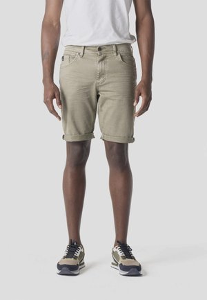 Hombre con pantalones cortos beige remangados y zapatillas beige con cordones negros, de pie contra un fondo liso gris claro.