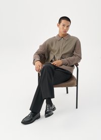 Ανδρικό μπουφάν με φερμουάρ σε γήινες αποχρώσεις, με υφή και υπερμέγεθεις τσέπες. Στενές μαύρες παντελόνες και γυαλιστερά μαύρα loafers.