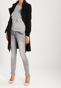 Femme portant un pull gris clair à volants, un jean skinny gris délavé, un manteau noir ouvert, et des chaussures à talons hauts roses, debout sur un fond blanc.