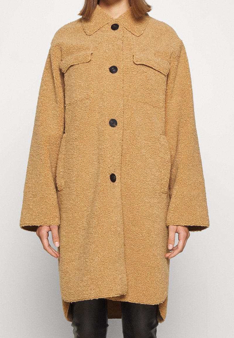 Cappotto in fleece beige con superficie testurizzata, grandi tasche sul petto, bottoni neri e orlo leggermente curvo. Maniche lunghe e larghe.