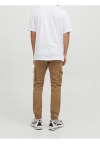 Braune Cargo-Hosen mit Seitentaschen, kombiniert mit einem schlichten weißen T-Shirt. Die Hose hat einen schmal zulaufenden Schnitt und elastische Knöchelabschlüsse. Weiße Sneaker sind sichtbar.