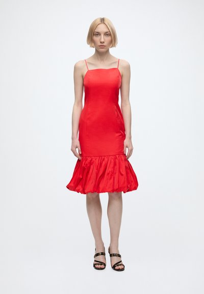 Femme aux cheveux blonds courts portant une robe rouge sans manches avec un ourlet à volants et des sandales noires à talons hauts à lanières, debout sur un fond blanc.