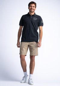 Czarny polo z kołnierzykiem i logo, w zestawieniu z beżowymi szortami. Model nosi białe trampki i skarpetki do kostek. Prosty i casualowy wygląd.