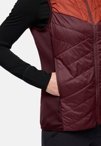 Donkerrode vest met een gewatteerde textuur aan de voorkant, contrasterend schoudergedeelte in een lichtere rode tint, en een zwart longsleeve shirt eronder.