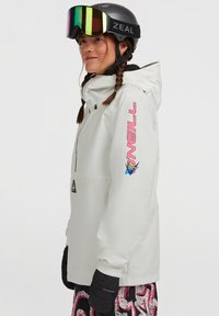 Witte snowboardjas met capuchon, voorzien van een roze logo op de mouw en een ritszak. Zwarte handschoen en meerkleuren broek zichtbaar eronder.
