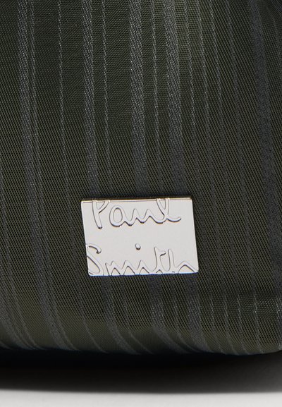 Svart stripete tekstilveske med et rektangulært sølvmerke preget med "Paul Smith" i håndskrift. Teksturen er glatt med subtile mønstre.