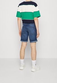 Persona vistiendo una camiseta a rayas en verde, blanco y azul marino, con pantalones cortos de denim azul remangados, calcetines blancos y zapatillas blancas, de pie de espaldas.