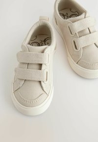 Beige canvas sneakers met twee klittenbandsluitingen, afgeronde neus, gestructureerde rubberen zool en een eenvoudig ontwerp. Het interieur heeft een gemusterde inlegzool.