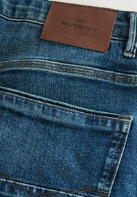 Modré džínsy z denimu s hnedou koženou nálepkou "DENIM SUPPLY" a dvojitým prešitím; majú zadnú vrecko a vyblednutú textúru.