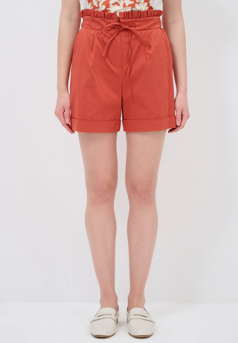 Pantalones cortos de color óxido con un ajuste relajado, cinturilla elástica y lazo delantero. La tela es suave y el dobladillo es vuelto. Combinados con zapatos blancos tipo slip-on.