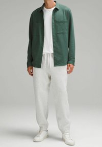 Camisa de botonadura larga de color verde sobre una camiseta blanca, combinada con pantalones de chándal gris claro y zapatillas blancas. Ajuste relajado y texturas suaves.