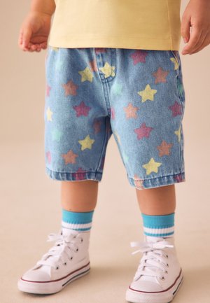 Barn iført blå shorts med flerfarvede stjerner, en gul skjorte, blå stribede sokker og hvide høje sneakers står på et lyst gulv.