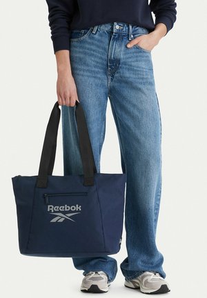 Osoba ubrana w niebieskie dżinsy i białe trampki, trzymająca granatową torbę Reebok z czarnymi uchwytami.