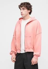 Ροζ κομμένο hoodie με φερμουάρ και κουκούλα, μπροστινές τσέπες και ακατέργαστες άκρες. Φορεμένο πάνω από λευκό πουκάμισο και συνδυασμένο με σκούρο παντελόνι.