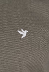 Tela gris texturizada con un logo de colibrí bordado en blanco en la esquina inferior. El material parece transpirable con un patrón similar a una malla.