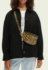 Veste noire avec des détails à franges, accompagnée d'un petit sac bandoulière zippé avec un motif rayé tigre jaune et noir.