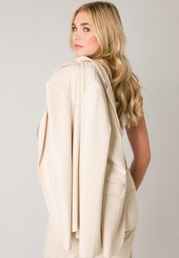 Beige blazer met gestructureerde schouders, één knoopsluiting en gladde stof, over de schouder van het model gedrapeerd tegen een neutrale achtergrond.