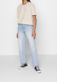 T-shirt beige in cotone con un piccolo logo bianco, abbinato a jeans dritti blu chiaro e sneakers alte nere con suole bianche.