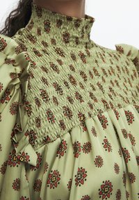 Blusa verde con motivo floreale, dotata di busto arricciato, maniche a volant e colletto alto. Presenta elementi di design in rosso e nero.