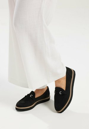 Zwarte instaploafers met bruine bies en zilveren gespsluiting, gedragen bij witte broek met wijde pijpen op een gladde, lichte achtergrond.