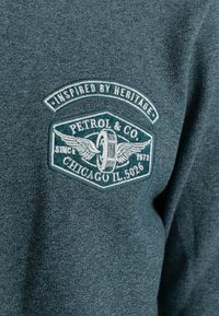 Türkisfarbener Pullover mit gesticktem Logo mit Flügeln, auf dem "Petrol & Co" und "Chicago IL 5026" zu lesen ist, auf einem strukturierten Stoff.