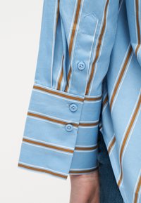 Μανσέτα μακριάς μπλούζας ανοιχτού γαλάζιου χρώματος με καφέ και λευκές κάθετες ρίγες, κουμπωμένη με δύο μπλε κουμπιά, φορεμένη από ένα άτομο με ορατό το κάτω μέρος του βραχίονα.