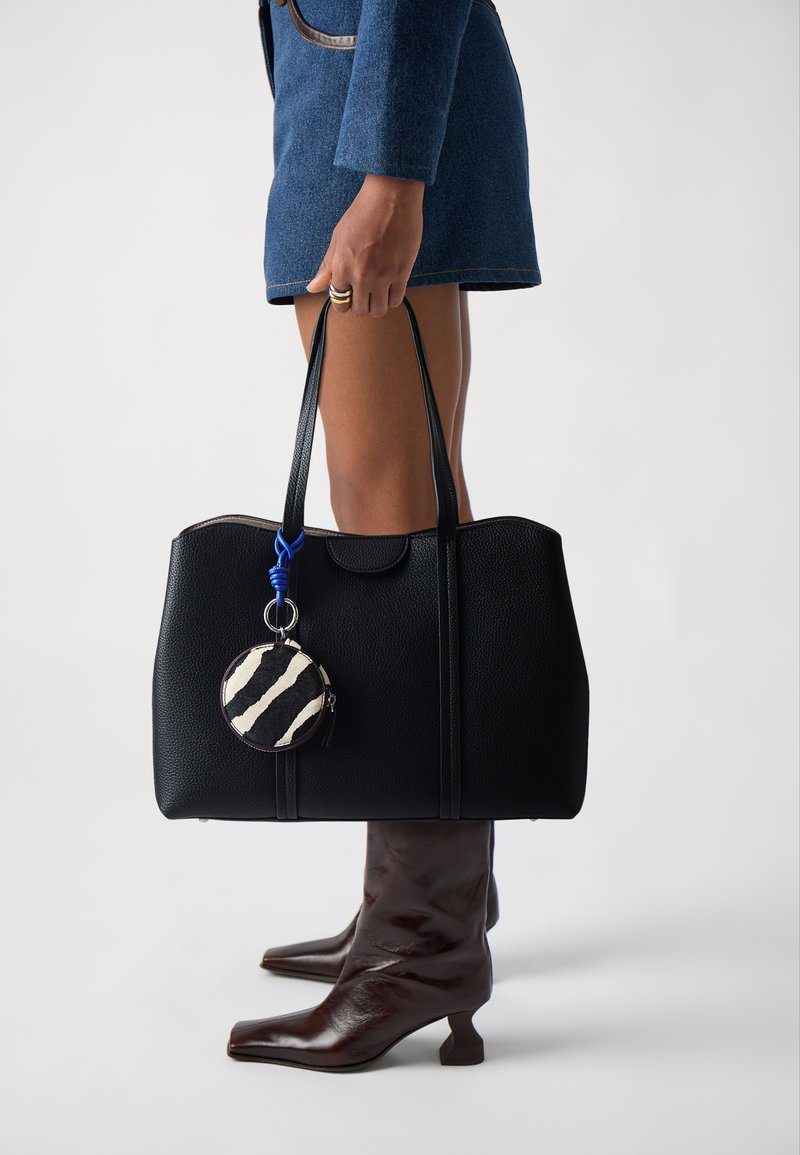 Borsa a tracolla nera in pelle con finitura testurizzata, caratterizzata da un portachiavi rotondo rigato in bianco e nero. Il modello indossa stivaletti marroni con tacco.