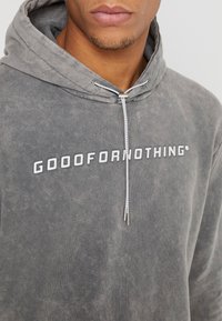 Grijze hoodie met een gestructureerde afwerking, voorzien van de tekst "GOOOFORNOTHING" in wit, uitgerust met een trekkoord en metalen uiteinden.