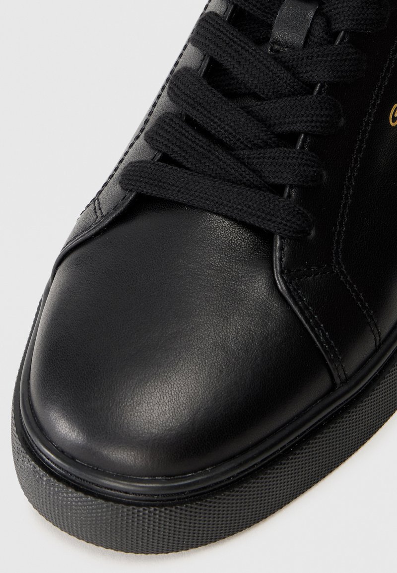Czarny skórzany sneaker z płaskimi czarnymi sznurówkami, zaokrąglonym noskiem, teksturowaną gumową podeszwą i minimalnymi detalami przeszycia.