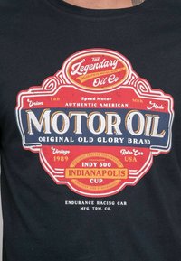 T-shirt noir avec un graphique de style vintage comprenant un design d'étiquette rouge et bleue, indiquant "Huile Moteur" et "Coupe d'Indianapolis", avec des accents vieillis.