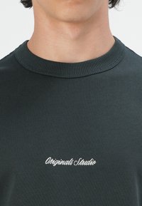 Camiseta de algodón verde oscuro con un cuello acanalado y texto bordado en blanco "Originals Studio" en el pecho. Textura suave y lisa.