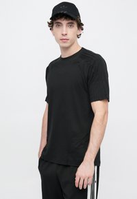 Μαύρο αθλητικό μπλουζάκι με κοντά μανίκια, με υφασμένα ραφές και εφαρμοστή γραμμή. Φορεμένο με μαύρο παντελόνι και καπέλο που ταιριάζει.