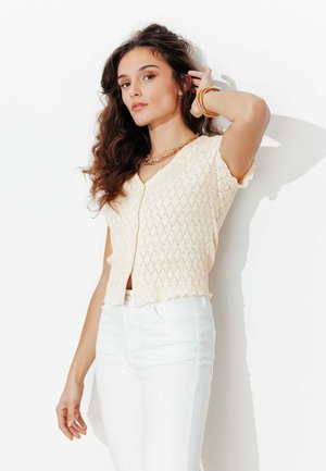 Femme aux cheveux bruns ondulés portant un haut en tricot crème à manches courtes et un pantalon blanc, accessoirisée de bracelets dorés et d'un collier.