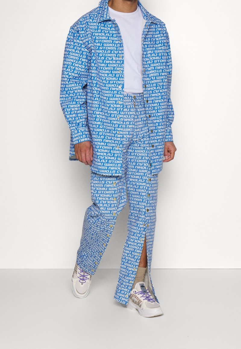 Homme portant une chemise bleue à motifs boutonnée et un pantalon assorti par-dessus un t-shirt blanc et des baskets blanches avec des lacets violets en train de marcher.